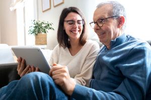 The Lodge at Stephens Lake | Happy senior man and his young daughter laughing together using digital tablet sitting on sofa at home